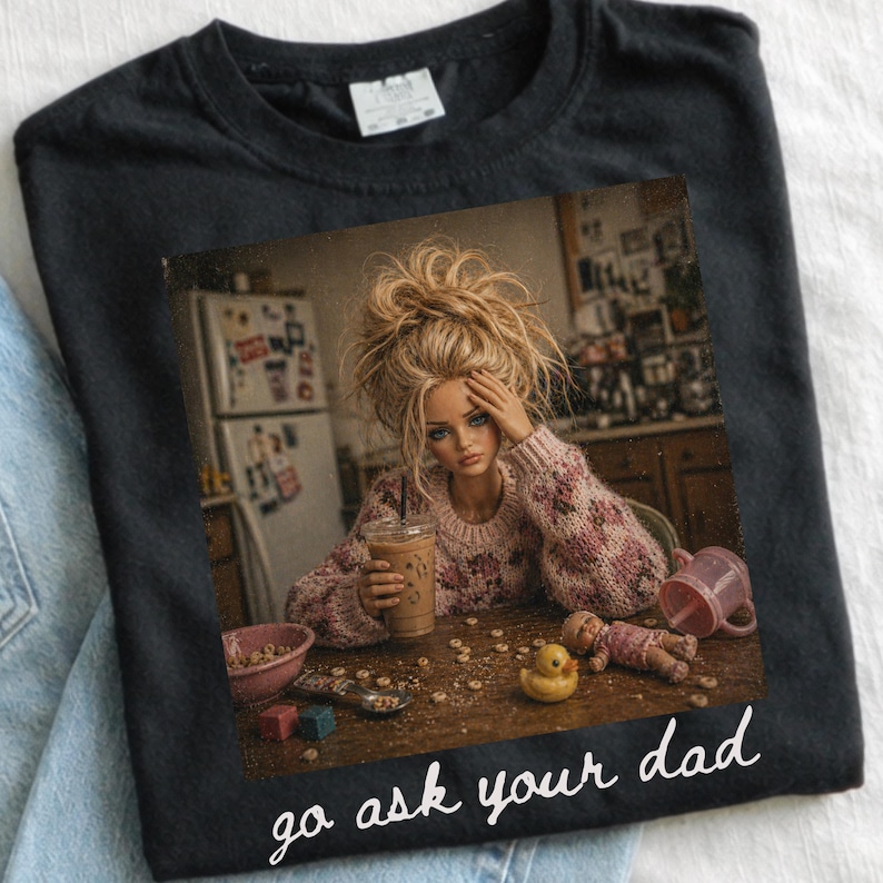 Pu&ograve; includere: T-shirt nero con un'immagine di una bambola che tiene una bevanda al caff&egrave;, circondata da giocattoli e cereali. Il testo "go ask your dad" &egrave; stampato in bianco sul fondo della maglietta.