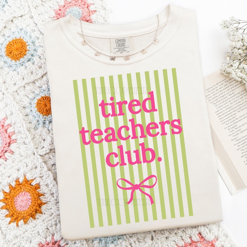 May include: Cream-colored sweatshirt with the text "tired teachers club." in pink letters, set against a background of green vertical stripes. A pink bow is below the text. A delicate butterfly necklace rests on the shirt.