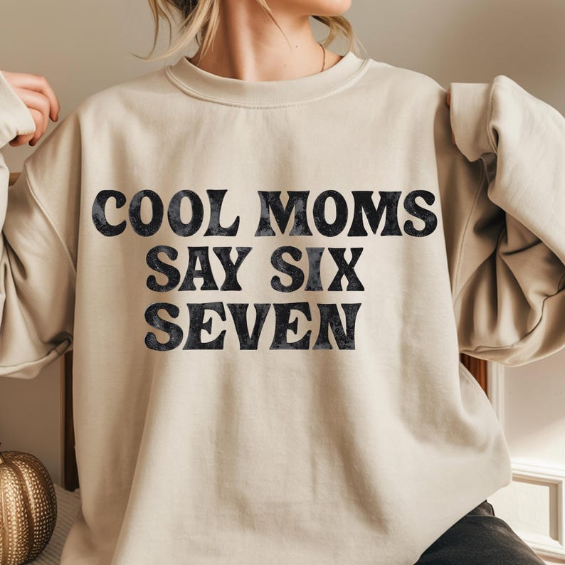 May include: A beige sweatshirt with the text "COOL MOMS SAY SIX SEVEN" in a dark, textured font. The crew neck sweatshirt is a neutral color and has long sleeves.