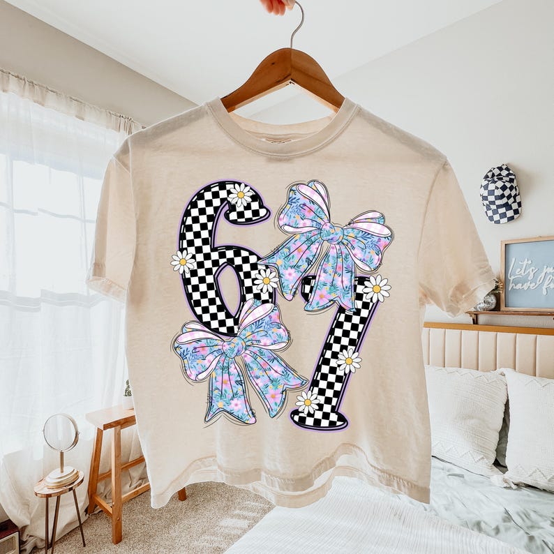May include: A beige t-shirt with a graphic design featuring the numbers "67" in a black and white checkered pattern. The numbers are adorned with floral bows and daisy accents. The shirt is hanging on a wooden hanger.