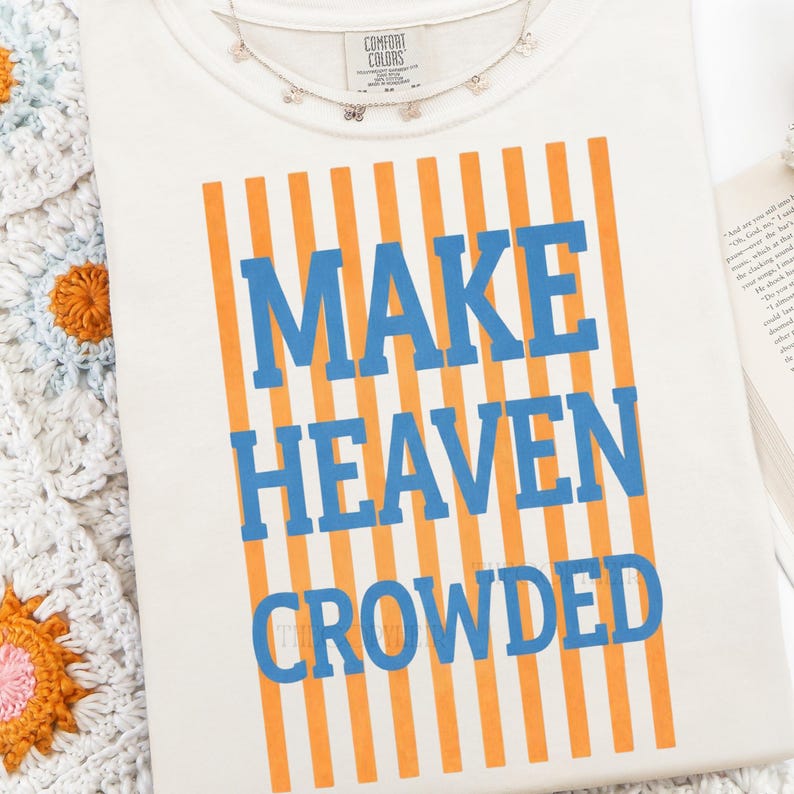 Puede incluir: Camiseta color crema con el texto "MAKE HEAVEN CROWDED" en letras azules, sobre rayas verticales naranjas. Un delicado collar de mariposas descansa sobre la camiseta. La camiseta est&aacute; sobre una superficie blanca.