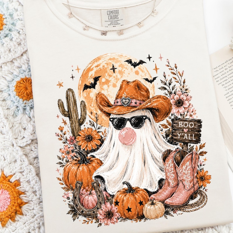 May include: A cream-colored t-shirt featuring a graphic of a ghost wearing a brown cowboy hat and black sunglasses, blowing a pink bubble. The design includes a full moon, bats, cacti, pumpkins, a horseshoe, and pink cowboy boots with text reading BOO Y'ALL.