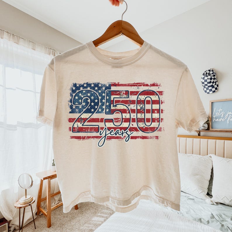 Puede incluir: Una camiseta color crema con un dise&ntilde;o desgastado de la bandera estadounidense. El gr&aacute;fico de la bandera presenta el n&uacute;mero "250" en azul y rojo, con la palabra "years" debajo. La camiseta est&aacute; colgada en una percha de madera.