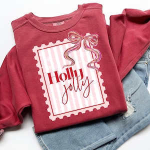 Puede incluir: Una sudadera granate con un dise&ntilde;o gr&aacute;fico a rayas rosas y blancas, un lazo decorativo y las palabras "Holly Jolly" en escritura roja. La sudadera est&aacute; doblada sobre unos pantalones cortos vaqueros azules.