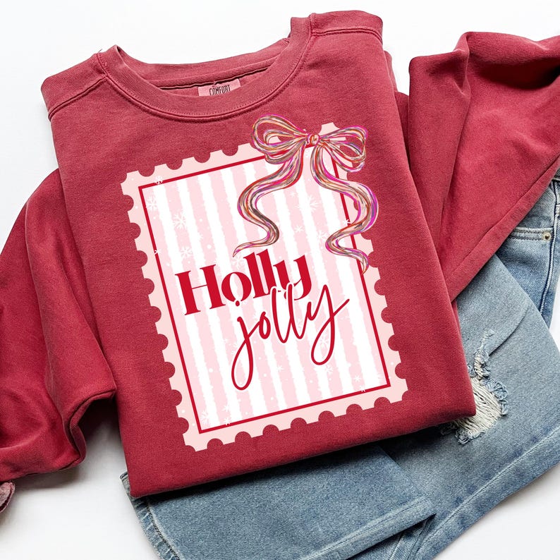 Puede incluir: Una sudadera granate con un dise&ntilde;o gr&aacute;fico a rayas rosas y blancas, un lazo decorativo y las palabras "Holly Jolly" en escritura roja. La sudadera est&aacute; doblada sobre unos pantalones cortos vaqueros azules.