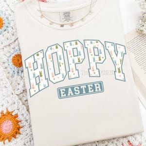 May include: Cream-colored sweatshirt with the words "HOPPY EASTER" in a blue and white collegiate-style font. The letters are filled with a pattern of pastel-colored Easter bunnies. A delicate necklace rests on the shirt.