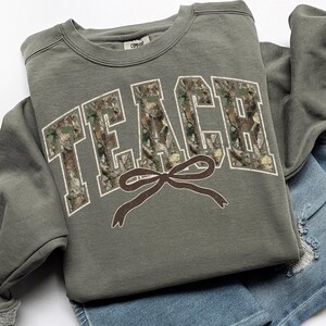May include: An olive green sweatshirt with the word "TEACH" in large, block letters filled with a camouflage pattern. A brown bow graphic is below the word. The sweatshirt is folded on top of a pair of blue denim shorts.