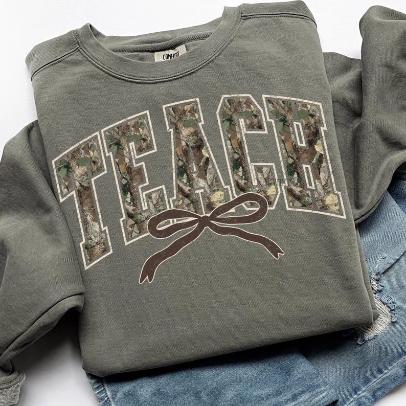 May include: An olive green sweatshirt with the word "TEACH" in large, block letters filled with a camouflage pattern. A brown bow graphic is below the word. The sweatshirt is folded on top of a pair of blue denim shorts.
