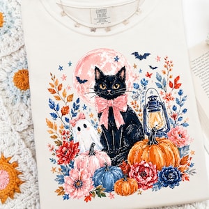 May include: A cream-colored t-shirt featuring a graphic print of a black cat wearing a pink bow, a ghost, a lantern, pumpkins, and flowers set against a pink moon. The shirt has a Comfort Colors label at the neckline and a crew neck design.