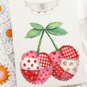 May include: Cream-colored t-shirt featuring a patchwork cherry design. The cherries are made of various patterned fabrics, including gingham, floral, and strawberry prints. The shirt has a crew neck and short sleeves. A delicate necklace with small charms is visible.