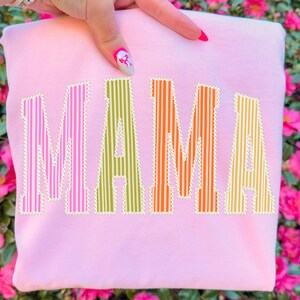 Puede incluir: Una sudadera rosa claro con la palabra "MAMA" en letras grandes, coloridas y a rayas. Las letras est&aacute;n delineadas en blanco con un patr&oacute;n de puntos. El fondo es un borr&oacute;n de flores rosas.