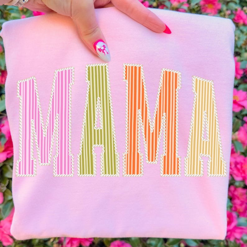 Puede incluir: Una sudadera rosa claro con la palabra "MAMA" en letras grandes, coloridas y a rayas. Las letras est&aacute;n delineadas en blanco con un patr&oacute;n de puntos. El fondo es un borr&oacute;n de flores rosas.