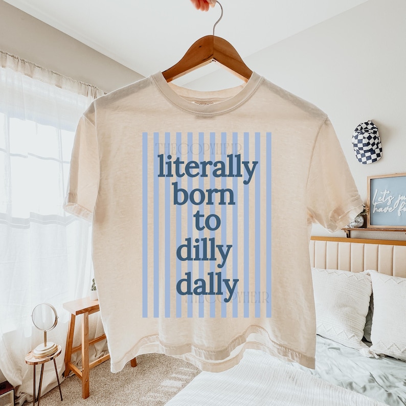 Puede incluir: Camiseta color crema con el texto "literally born to dilly dally" en azul. El texto est&aacute; centrado en un fondo a rayas azul claro. La camiseta est&aacute; colgada en una percha de madera.