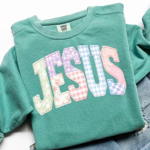Puede incluir: Una sudadera Comfort Colors verde azulado con la palabra "JESUS" aplicada en la parte delantera. Las letras son de patchwork con estampados florales, cuadros y lisos en colores pastel. La sudadera est&aacute; doblada sobre una superficie blanca.