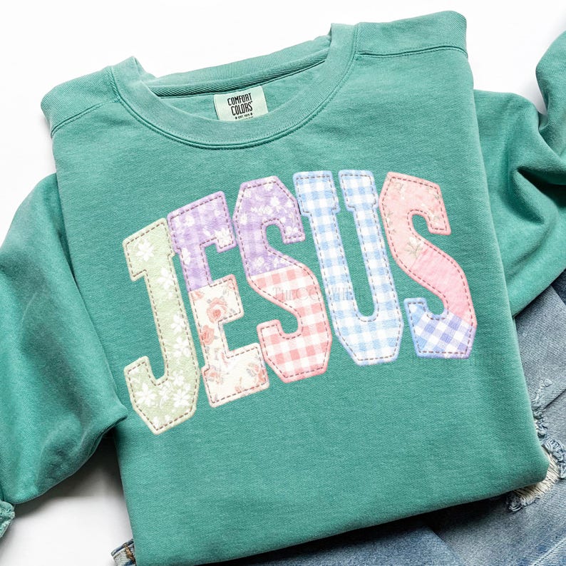 Puede incluir: Una sudadera Comfort Colors verde azulado con la palabra "JESUS" aplicada en la parte delantera. Las letras son de patchwork con estampados florales, cuadros y lisos en colores pastel. La sudadera est&aacute; doblada sobre una superficie blanca.