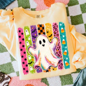 May include: A pale yellow crewneck sweatshirt featuring a large central graphic of a white ghost. The design includes vertical brushstroke stripes in pink, green, purple, orange, and teal, decorated with ghosts, bats, spiders, and candy corn.