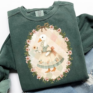 May include: Dark green sweatshirt featuring a whimsical illustration of a goose in a dress holding a flower, framed by a floral wreath. The design includes a bow and the text "THECOOPYHEIR". The sweatshirt has a crew neck and long sleeves.