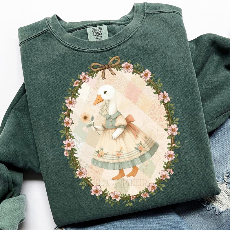 May include: Dark green sweatshirt featuring a whimsical illustration of a goose in a dress holding a flower, framed by a floral wreath. The design includes a bow and the text "THECOOPYHEIR". The sweatshirt has a crew neck and long sleeves.