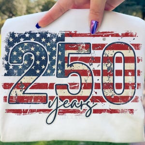 Puede incluir: Camiseta blanca con un dise&ntilde;o desgastado de la bandera estadounidense. Los n&uacute;meros "250" se muestran de forma destacada, con la palabra "years" en escritura cursiva debajo. El dise&ntilde;o utiliza los colores rojo, blanco y azul, evocando un tema patri&oacute;tico.