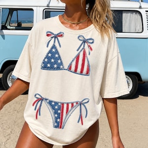Op de afbeelding: Crèmekleurig T-shirt met een patriottisch bikiniontwerp. De bikinitop heeft een blauwe denim driehoek met witte sterren en een rood-wit gestreepte driehoek. De bijpassende broek heeft hetzelfde ontwerp. Het shirt heeft een relaxte pasvorm.