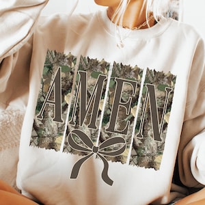 K&ouml;nnte beinhalten: Cremefarbenes Sweatshirt mit dem Wort "AMEN" in brauner Schrift, vor einem Camouflage-Hintergrund. Unter dem Wort befindet sich eine braune Schleife. Das Sweatshirt ist ein l&auml;ssiger, bequemer Stil.