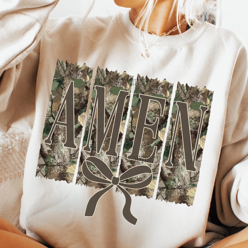 K&ouml;nnte beinhalten: Cremefarbenes Sweatshirt mit dem Wort "AMEN" in brauner Schrift, vor einem Camouflage-Hintergrund. Unter dem Wort befindet sich eine braune Schleife. Das Sweatshirt ist ein l&auml;ssiger, bequemer Stil.