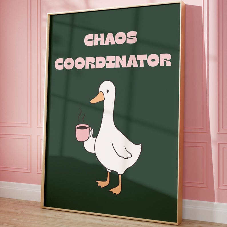 May include: A framed art print with a white cartoon goose holding a pink coffee mug. The words "CHAOS COORDINATOR" are printed in pink above the goose on a dark green background. The frame is gold.