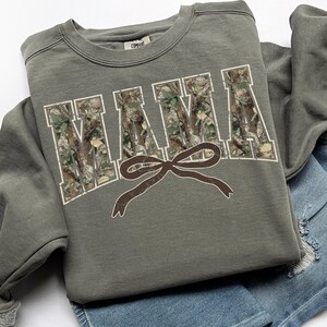 May include: An olive green sweatshirt with the word "MAMA" in large, white, block letters filled with a camouflage pattern. A brown bow is centered below the word. The sweatshirt is folded on top of a pair of blue denim shorts.
