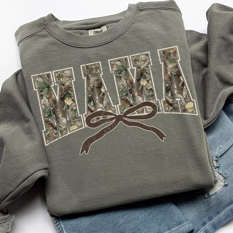 May include: An olive green sweatshirt with the word "MAMA" in large, white, block letters filled with a camouflage pattern. A brown bow is centered below the word. The sweatshirt is folded on top of a pair of blue denim shorts.