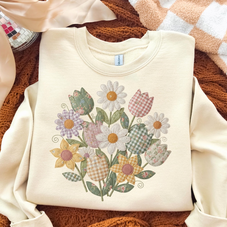 May include: Cream-coloured sweatshirt with a floral design. The design features daisies and tulips in a patchwork style. The flowers are in shades of yellow, pink, green, and purple. The sweatshirt has a crew neck and long sleeves.