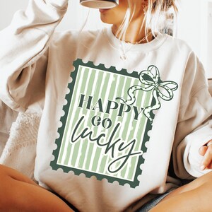 Puede incluir: Sudadera color crema con un gr&aacute;fico verde y blanco. El gr&aacute;fico presenta un dise&ntilde;o de sello postal con las palabras "Happy Go Lucky" y un lazo con acentos de tr&eacute;bol. La sudadera es de estilo casual y c&oacute;modo.