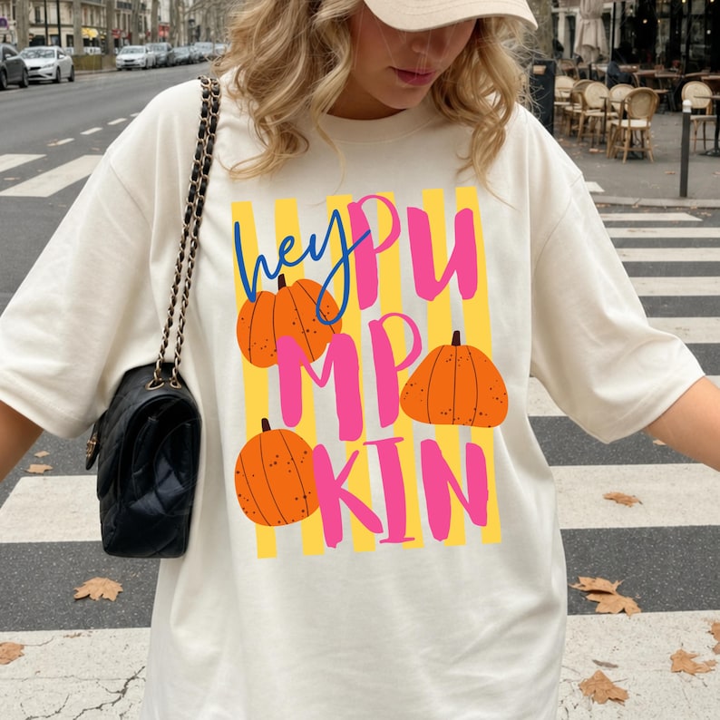 May include: Cream-colored t-shirt with the text "hey PUMPKIN" in blue, pink, and yellow. The design includes orange pumpkin illustrations. The shirt is a casual, oversized fit.