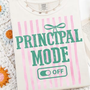 May include: Cream-colored t-shirt with pink vertical stripes and the words "PRINCIPAL MODE OFF" in green. A green bow graphic is above the word "PRINCIPAL". A delicate butterfly necklace is at the neckline.