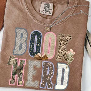 May include: A brown Comfort Colors t-shirt with the words "BOOK NERD" in a collage style. The letters are decorated with floral, leopard print, and book page patterns. A silver necklace with a pendant is also visible.