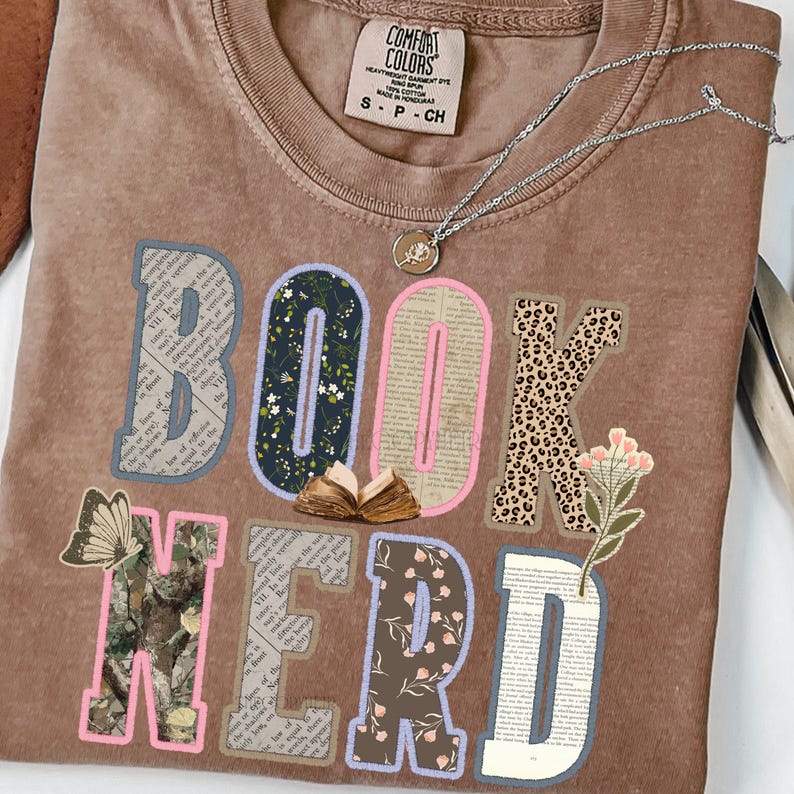 May include: A brown Comfort Colors t-shirt with the words "BOOK NERD" in a collage style. The letters are decorated with floral, leopard print, and book page patterns. A silver necklace with a pendant is also visible.