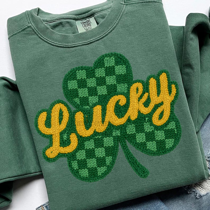 K&ouml;nnte beinhalten: Gr&uuml;nes Sweatshirt mit einem gro&szlig;en Kleeblatt-Design. Das Kleeblatt ist gr&uuml;n mit einem Karomuster, und das Wort "Lucky" ist in gelben, gestickten Buchstaben gehalten. Das Sweatshirt hat einen Rundhalsausschnitt und lange &Auml;rmel.