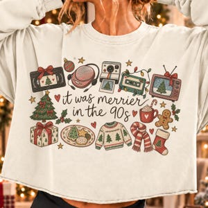 May include: A cream-colored crewneck sweatshirt featuring a colorful graphic print with the text "it was merrier in the 90s". The design includes illustrations of a VHS tape, headphones, a camera, a cassette, a television, a tree, and gifts.