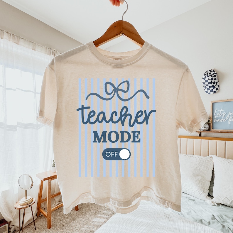 May include: Cream-colored, short-sleeved t-shirt with the words "teacher MODE OFF" in blue script and a bow graphic. The design includes light blue vertical stripes. The shirt is hanging on a wooden hanger.