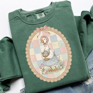 Puede incluir: Sudadera verde con un dise&ntilde;o ovalado de un ganso con gorro y vestido, llevando una cesta de huevos de Pascua. El dise&ntilde;o est&aacute; sobre un fondo de patchwork pastel con un lazo rosa en la parte superior.