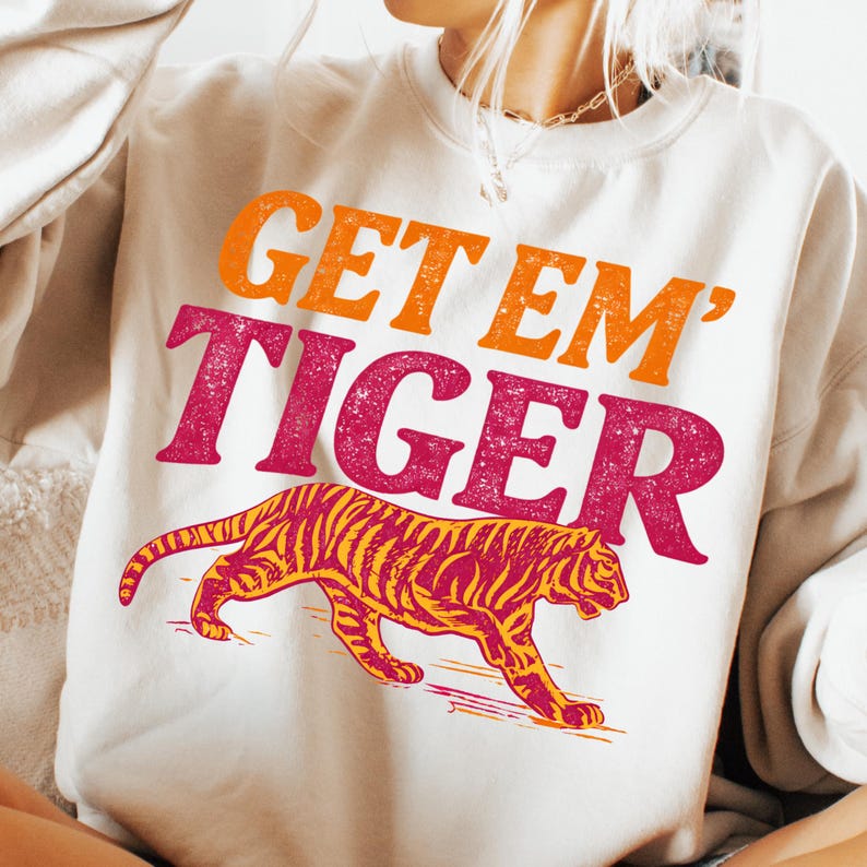 K&ouml;nnte beinhalten: Cremefarbenes Sweatshirt mit dem Text "GET EM' TIGER" in Orange und Fuchsia. Eine gelb-rote Tigergrafik befindet sich unter dem Text. Das Sweatshirt ist ein l&auml;ssiger Stil.