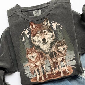 Puede incluir: Sudadera gris oscuro con una ilustraci&oacute;n detallada de tres lobos en un entorno forestal. Los lobos est&aacute;n representados en tonos marrones y grises, con un fondo de monta&ntilde;a y &aacute;rboles. La sudadera tiene cuello redondo y la etiqueta de la marca Comfort Colors.