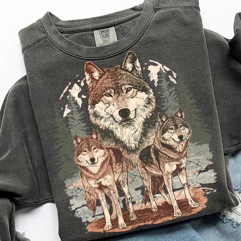 Puede incluir: Sudadera gris oscuro con una ilustraci&oacute;n detallada de tres lobos en un entorno forestal. Los lobos est&aacute;n representados en tonos marrones y grises, con un fondo de monta&ntilde;a y &aacute;rboles. La sudadera tiene cuello redondo y la etiqueta de la marca Comfort Colors.
