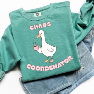 May include: Teal-colored sweatshirt with the words "CHAOS COORDINATOR" in pink. A cartoon goose holding a pink mug is in the center. The sweatshirt is folded on top of a pair of blue jeans.