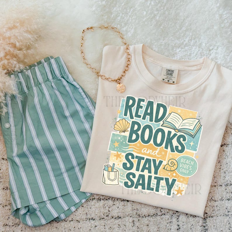以下が含まれることがあります： カラフルなビーチをテーマにしたデザインで「Read Books and Stay Salty」と書かれたクリーム色のTシャツ。画像には、緑と白のストライプのショートパンツと金のネックレスも含まれています。