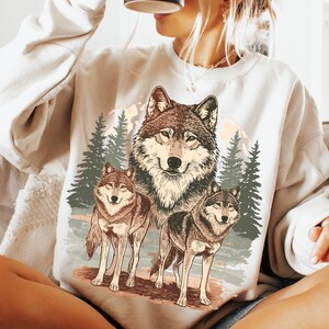Puede incluir: Sudadera de color crema con un gr&aacute;fico detallado de tres lobos en un entorno forestal. El dise&ntilde;o incluye un lobo central grande y dos lobos m&aacute;s peque&ntilde;os, con pinos y una cordillera en el fondo.