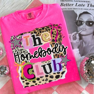 May include: A bright pink t-shirt with the words "The Homebody Club" in a collage-style design. The design includes leopard print, star accents, and colorful letters. The shirt is displayed on a pink and white checkered surface.
