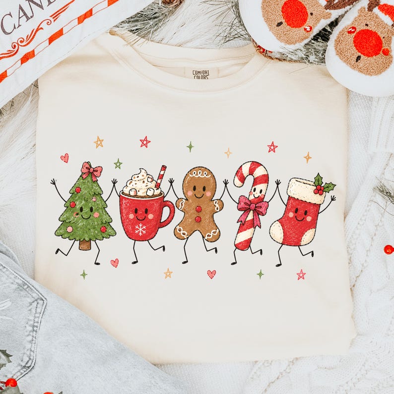 May include: A cream-colored crewneck t-shirt featuring a graphic print of five anthropomorphic Christmas characters holding hands: a green pine tree, a red mug of hot cocoa, a brown gingerbread man, a candy cane, and a red stocking.
