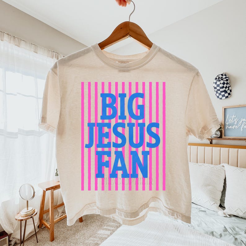 K&ouml;nnte beinhalten: Cremefarbenes T-Shirt mit dem blauen Text "BIG JESUS FAN" auf einem rosa gestreiften Hintergrund. Das Shirt h&auml;ngt an einem Holzb&uuml;gel.