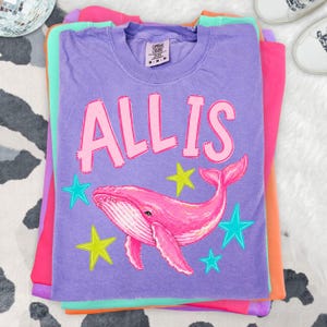 May include: A stack of colorful t-shirts, including purple, pink, and orange. The top shirt is purple and features the word "ALLIS" in pink letters above a pink whale illustration with teal and yellow stars. The shirts are folded.