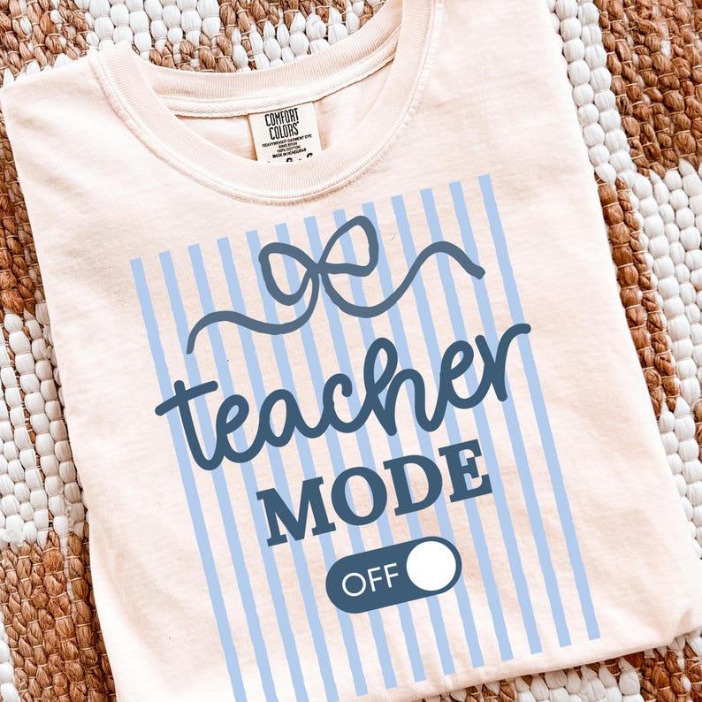 May include: A light peach-colored t-shirt with a blue design. The design includes a bow, vertical stripes, and the words "teacher MODE OFF" in a playful font. The shirt is made of a soft material.